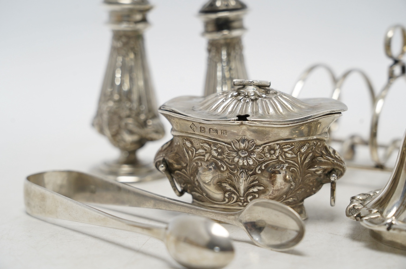 Sundry silver including a pair of Edwardian pepperettes, a five bar toast rack, a mustard pot (a.f.), a pair of sugar tongs and a cover. Condition - poor to fair